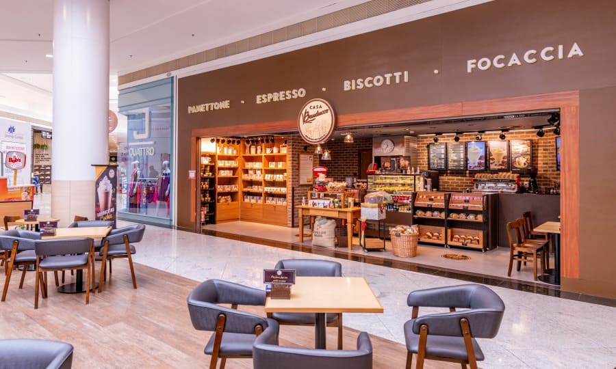 Imagem do interior do shopping, mostrando a loja Casa Bauducco com uma fachada de estilo rústico. A loja exibe prateleiras com produtos como panettones, biscoitos e focaccia. Na frente, mesas e cadeiras de madeira estão dispostas em um espaço aconchegante. O ambiente é iluminado e acolhedor, com a marca 'Casa Bauducco' visível na parte superior da loja