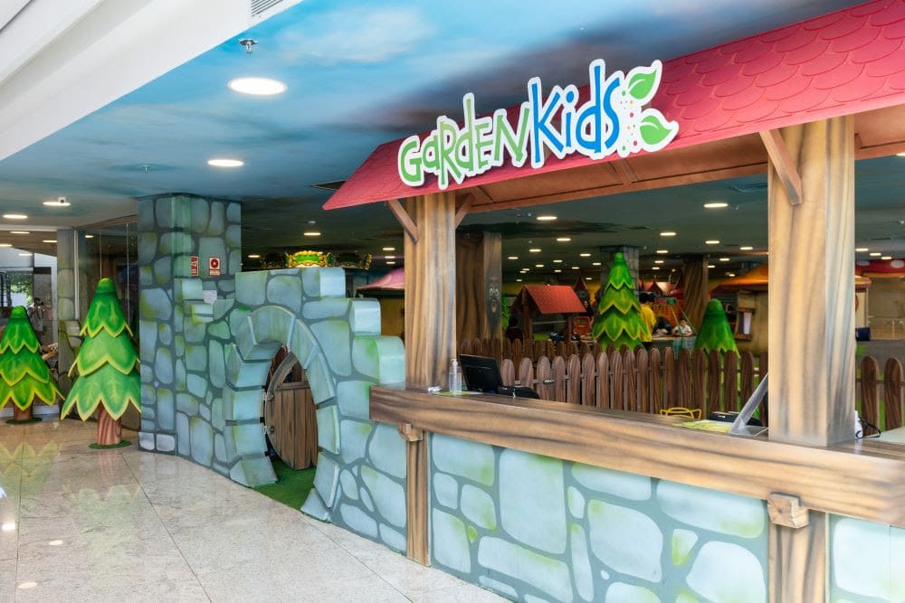 Imagem da entrada do espaço infantil 'Garden Kids' em um shopping, com decoração temática de jardim. A fachada é composta por elementos que imitam pedras e madeira, com um arco estilizado e uma placa colorida com o nome 'Garden Kids' no topo. Ao fundo, é possível ver árvores decorativas e estruturas lúdicas que compõem o ambiente interno para brincadeiras infantis.