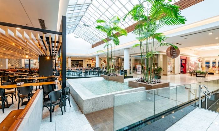 Interior de um shopping moderno com área de alimentação, mesas, plantas e uma fonte decorativa.