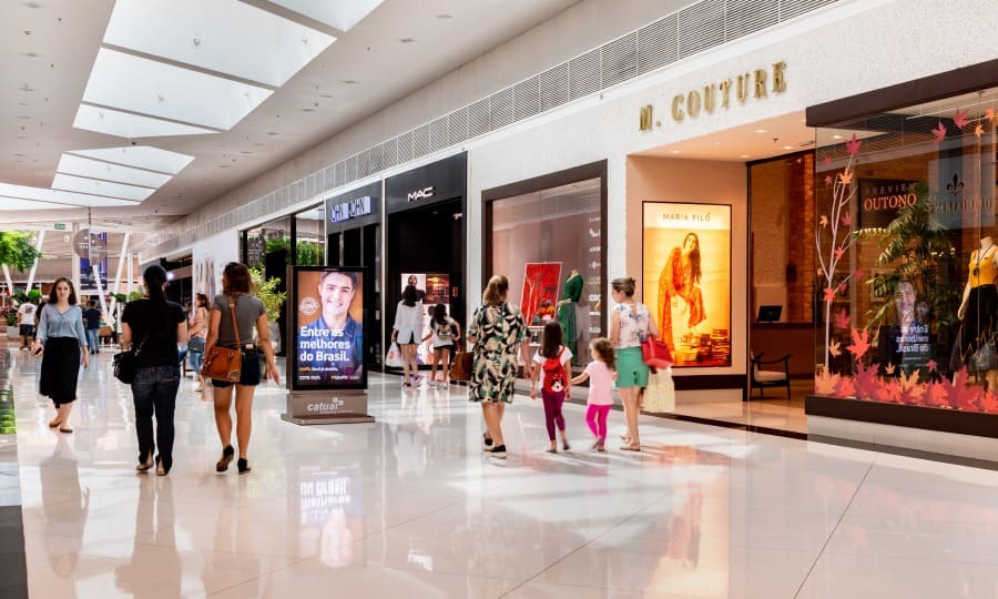 Foto interna do corredor com as lojas Catuaí Shopping, teto de vidro com iluminação natual e pessoas andando no local.