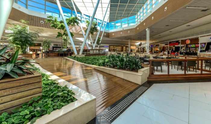 Interior de um shopping com áreas verdes, mesas e cadeiras, e iluminação natural.