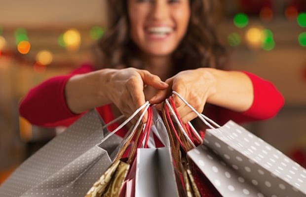 Imagem de uma mulher sorridente segurando várias sacolas de compras com aparência natalina. Ao fundo, há luzes desfocadas em tons quentes, criando um clima festivo e acolhedor. As sacolas possuem designs variados, incluindo padrões de bolinhas e detalhes dourados.