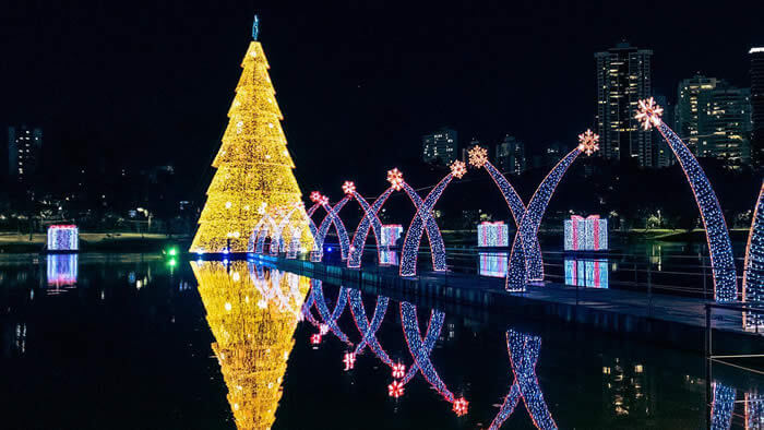 Árvore de Natal iluminada refletindo na água em uma noite clara. Decoração festiva com luzes brilhantes ao redor da lagoa, dando o intuito de economizar com passeios gratuitos.