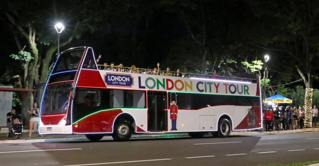 Ônibus do London City Tour iluminado, com passageiros no topo, passando por uma rua à noite rodeada de árvores e pessoas.
